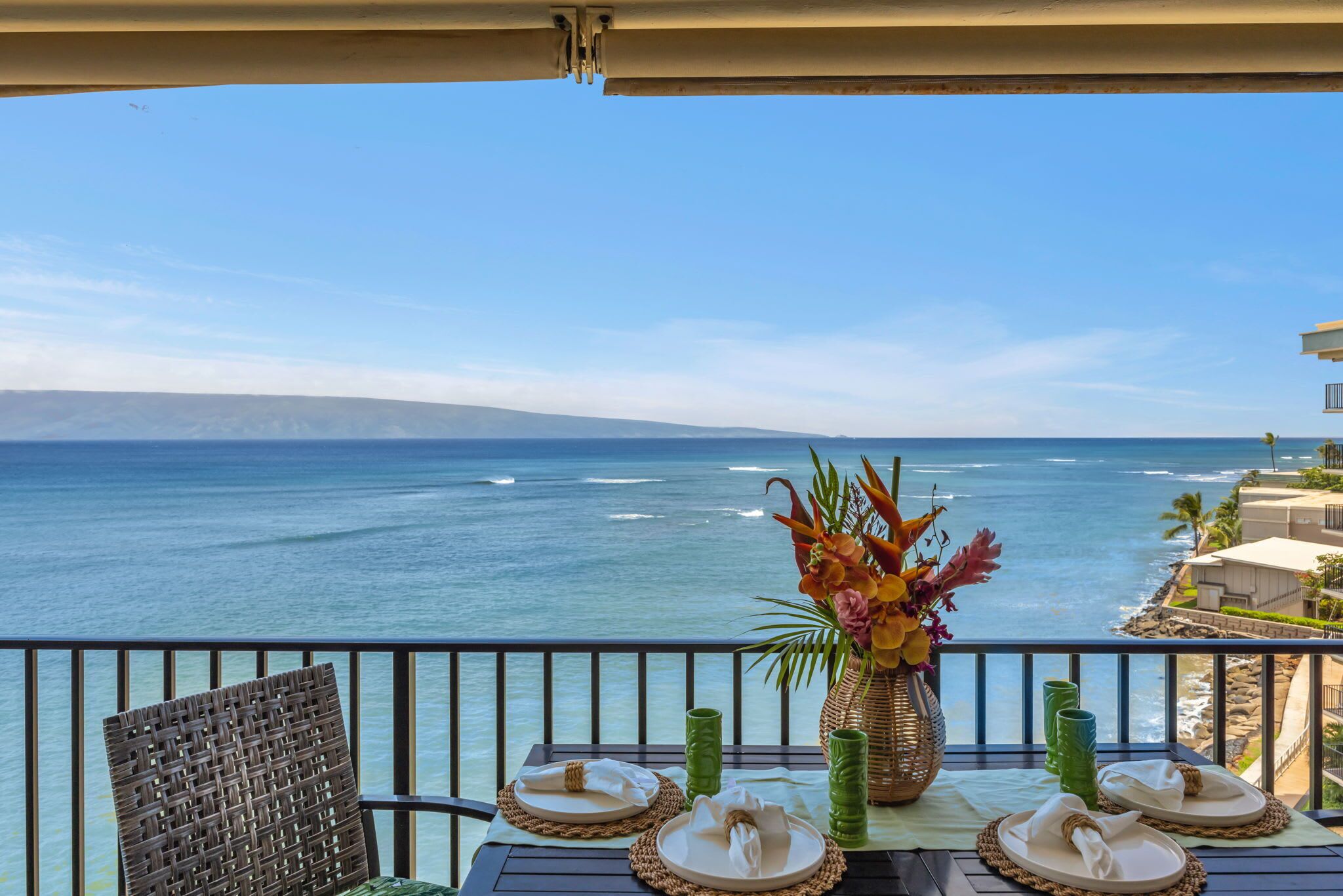 A dining table with plates, napkins, and a vase of flowers on a balcony overlooking the ocean.
