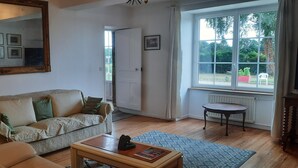 Een woonkamer met een bank, salontafel, stoel, spiegel, deur en raam met uitzicht op groen.