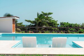 Une piscine avec deux chaises longues blanches, une clôture en bois et une végétation luxuriante en arrière-plan.