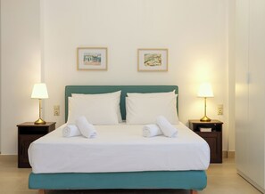 Un lit double bien fait, avec du linge de lit blanc et deux oreillers, flanqué de deux tables de chevet avec des lampes, dans une chambre dont la tête de lit est verte et où deux tableaux encadrés ornent le mur.