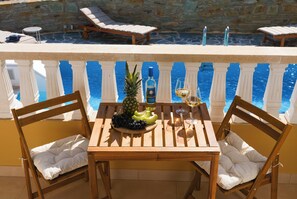 Une table en bois avec des chaises, une bouteille d’alcool, un ananas et un verre de vin sur un balcon, avec une piscine en arrière-plan.