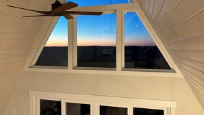Une pièce avec un plafond en pente, un ventilateur de plafond et une vue sur le coucher de soleil à travers une grande fenêtre.