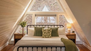 Une chambre avec un plafond en pente, un lit agrémenté de plusieurs oreillers, une table de chevet avec une lampe et une fenêtre donnant sur une zone boisée.