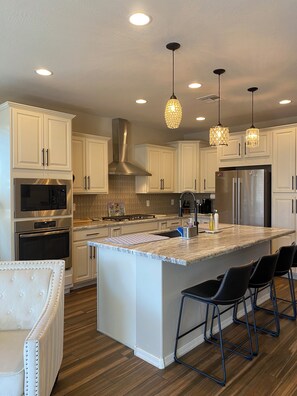 Cocina moderna con armarios blancos, electrodomésticos de acero inoxidable, una isla central con taburetes y luminarias colgantes.