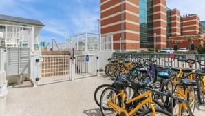 Un espace de stationnement pour vélos où sont garés plusieurs vélos, une entrée fermée par un portail et, en arrière-plan, un bâtiment moderne doté de fenêtres réfléchissantes.