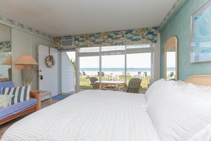Ein Schlafzimmer mit einem großen Bett, einem kleinen Tisch und Stühlen. Durch die Fenster ist der Strand und der Ozean zu sehen.