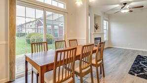 Een eetkamer met een grote tafel en stoelen, een plafondventilator en uitzicht op een huis door grote ramen.