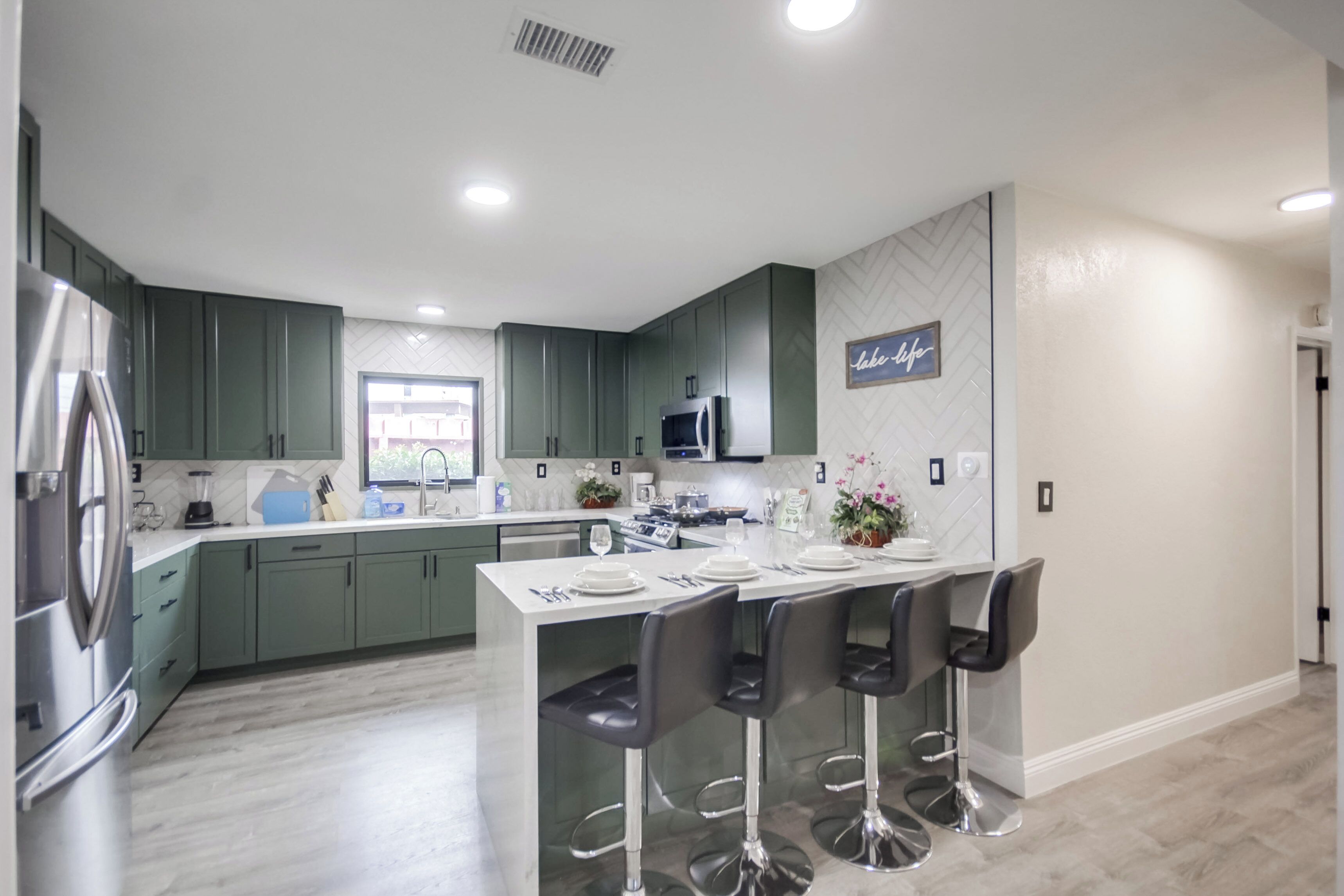Een moderne keuken met groene kastjes, een centraal eiland met barstoelen, roestvrijstalen apparatuur en een backsplash met een visgraatmotief.