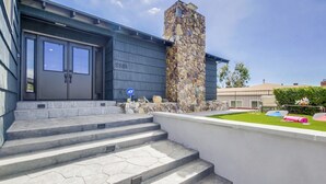 Un edificio moderno con una columna de piedra, una serie de escalones que conducen a una puerta y un pequeño jardín con sombrillas de colores.