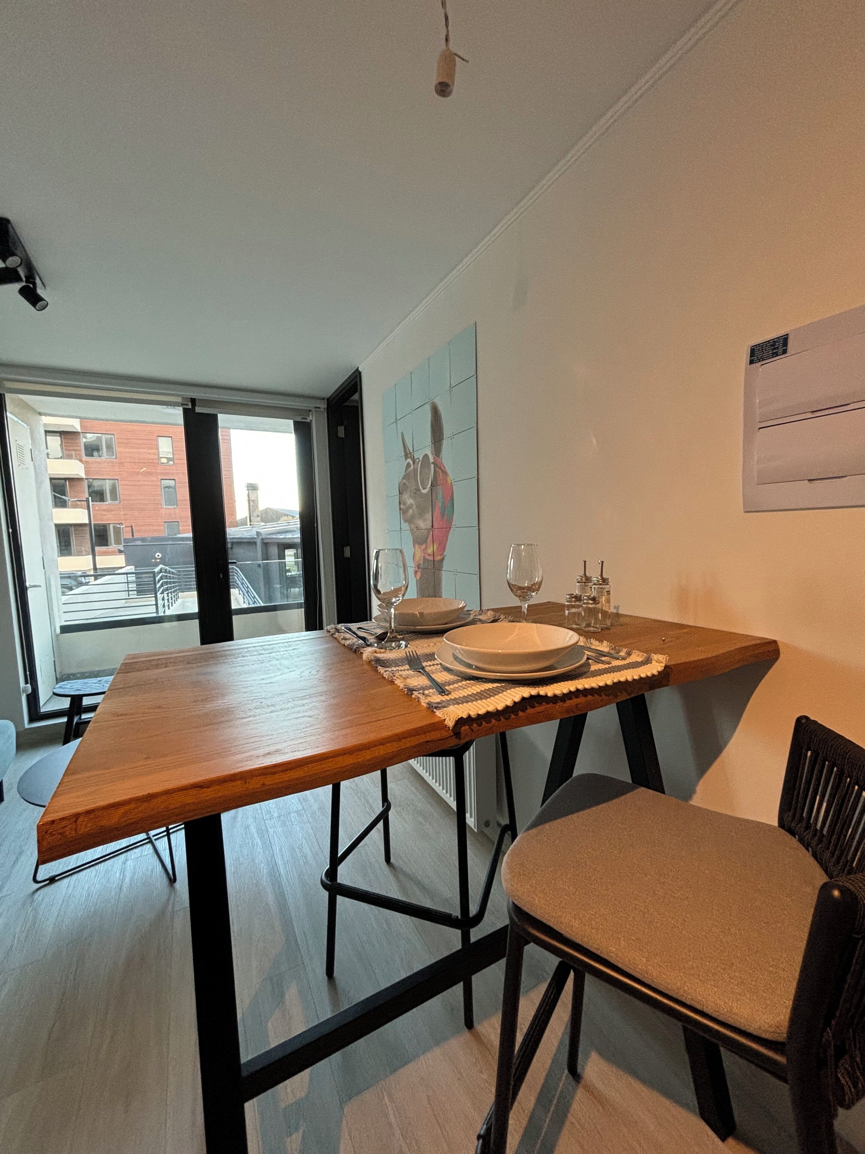 A dining area with a wooden table set for two, a chair, and a view of the city through a large window.