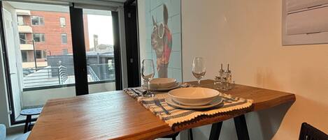 A dining area with a wooden table set for two, a chair, and a view of the city through a large window.