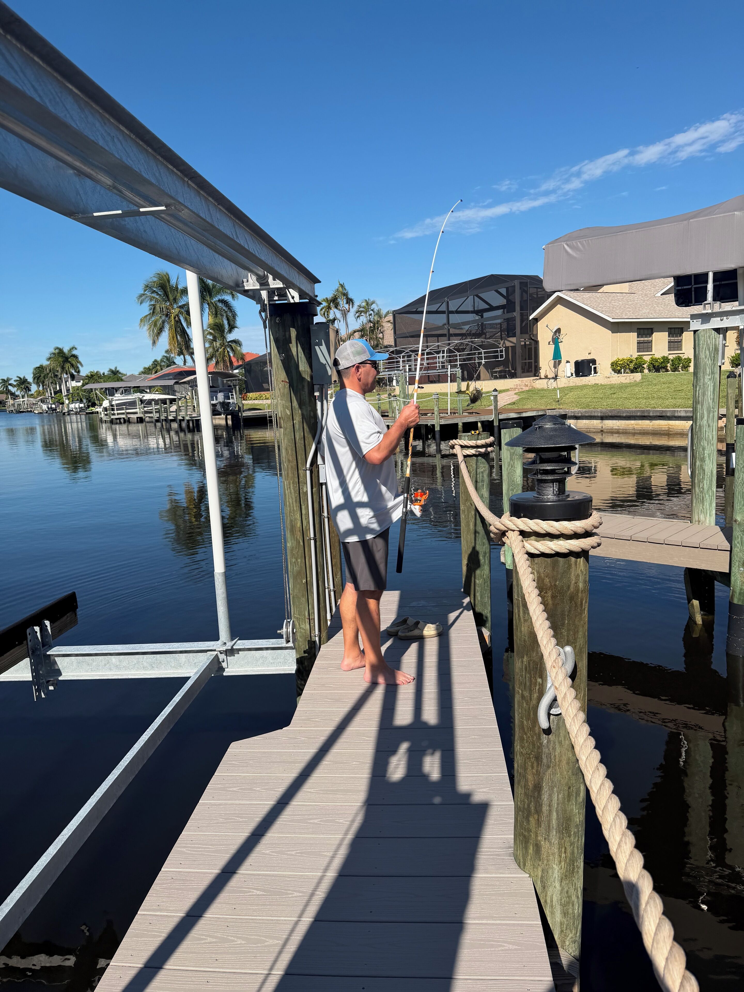 A man fishing on a wooden dock with a house and palm trees in the background.