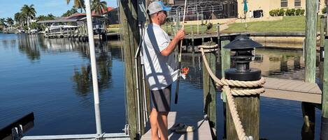 A man fishing on a wooden dock with a house and palm trees in the background.