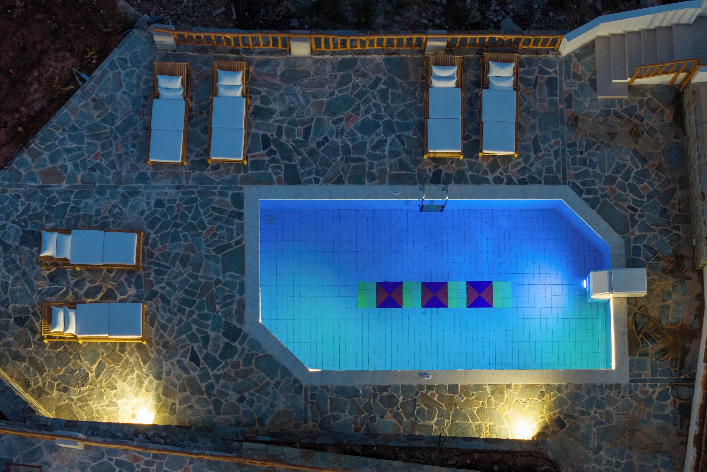 Vista aérea de una piscina con paredes de azulejos y piso de piedra, rodeada de sillones de descanso y una terraza de madera.