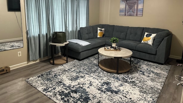 A living room with a gray sectional sofa, a round coffee table, a blue and white patterned rug, a ceiling fan, and a painting on the wall. - Soldier/Family Charming 3bed/2bath (Columbus)