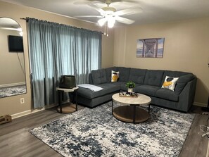 A living room with a gray sectional sofa, a round coffee table, a blue and white patterned rug, a ceiling fan, and a painting on the wall.