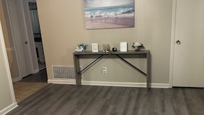 A room with a wooden floor, a large abstract rug, a console table with decorative items, a painting of a beach scene, and a door. - Soldier/Family Charming 3bed/2bath (Columbus)