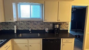 A kitchen with white cabinets, a black countertop, a window with blinds, a coffee maker, and a dishwasher. - Soldier/Family Charming 3bed/2bath (Columbus)