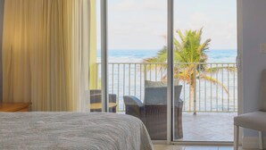 Habitación de hotel con cama, balcón y vista al océano y palmeras, y puerta corrediza de vidrio.