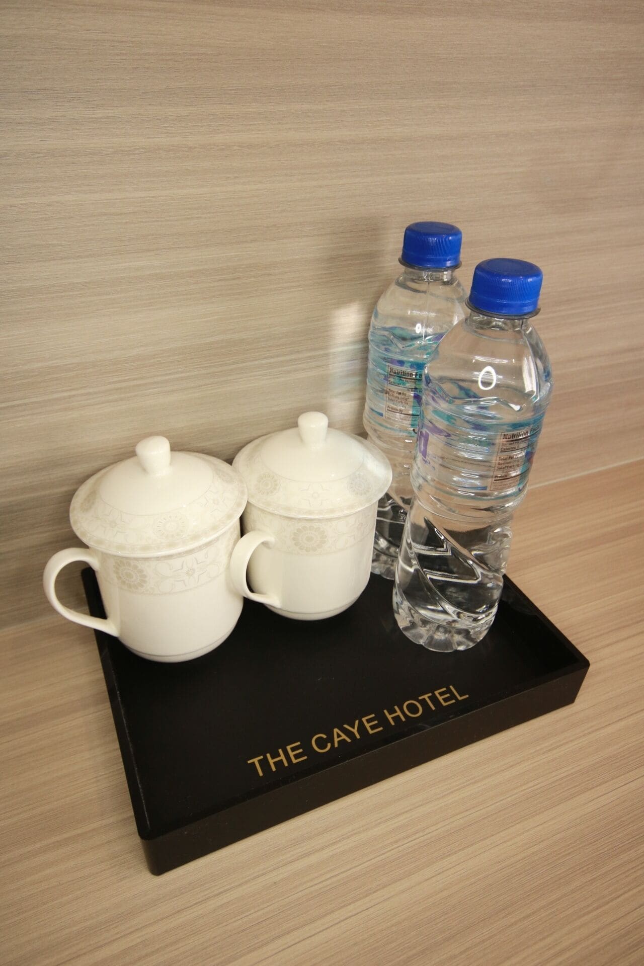 Un plateau avec deux tasses en céramique blanche et deux bouteilles d’eau avec des bouchons bleus, portant l’inscription « THE CAYE HOTEL ».