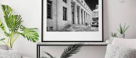 Une photographie en noir et blanc, encadrée, représentant un bâtiment à colonnes, est accrochée au-dessus d’une table ronde sur laquelle sont posés une plante et des livres. De part et d’autre de la table se trouvent deux fauteuils dotés de coussins moelleux.