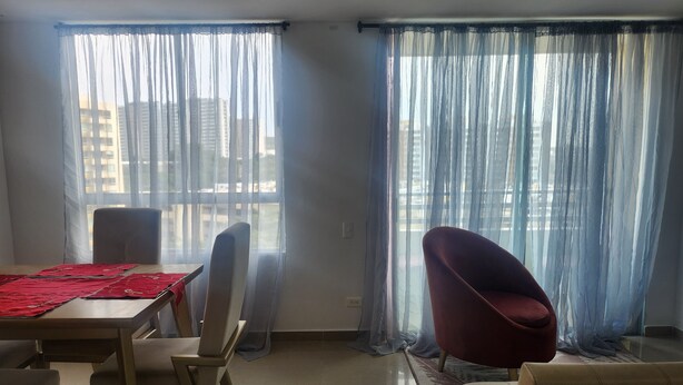 A living room with a dining area, large windows with sheer curtains, and a view of high-rise buildings. - Beautiful apartment to rest and vacation near puerto colombia. (Atlántico)