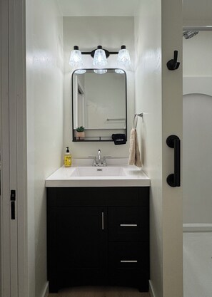 A bathroom with a white sink, black cabinets, a mirror with lights, a towel rack, and a door with a handle. - Cozy Riverfront Retreat (Suamico)