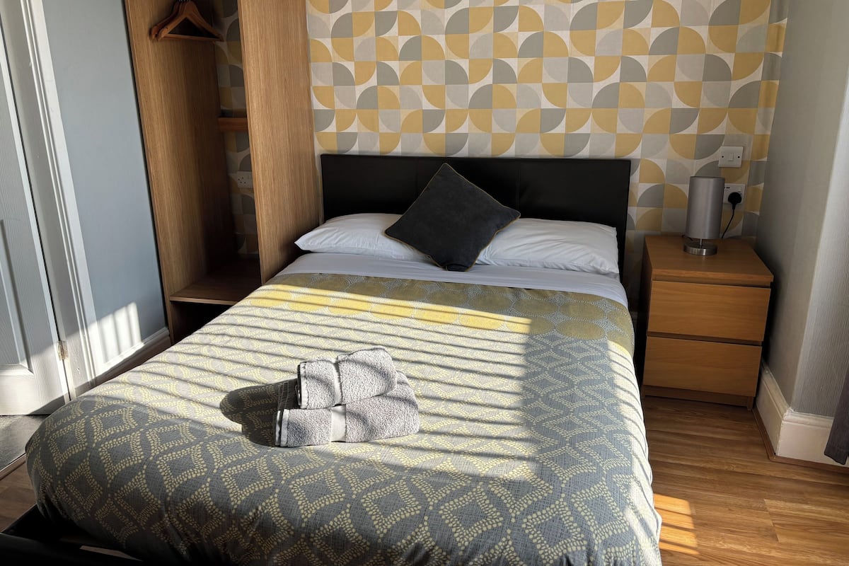 Una cama bien hecha con ropa de cama estampada, una mesita de noche con lámpara y un armario con ropa colgada.
