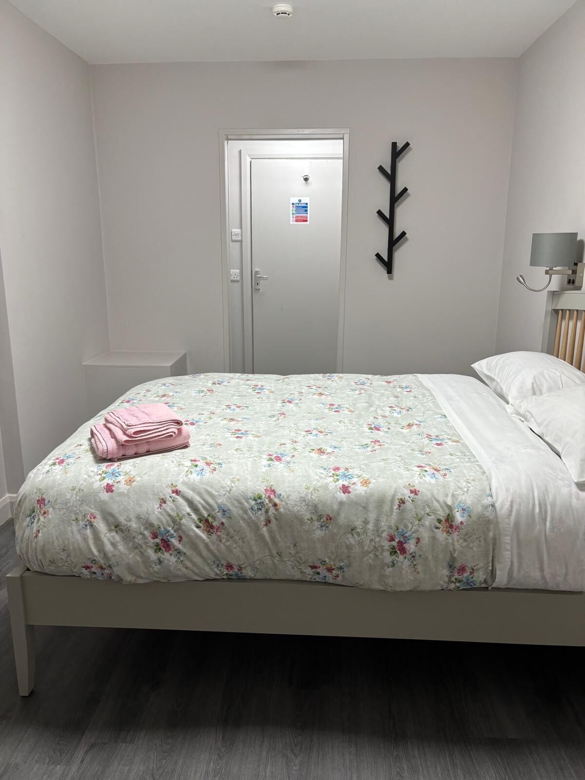 A bedroom with a bed, a door, a wall-mounted light, and a small shelf.