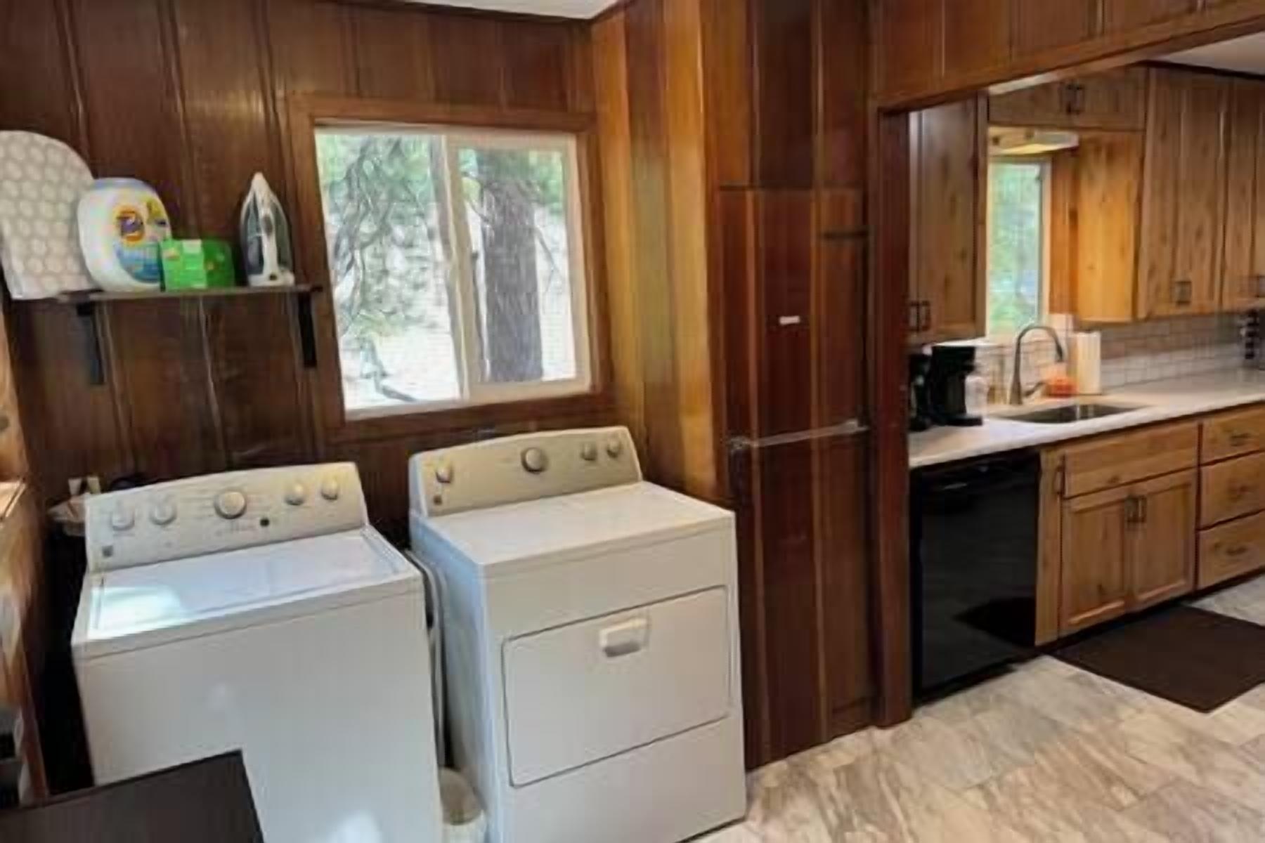 A laundry room with two white washing machines, wooden cabinets, and a window with a view of trees.