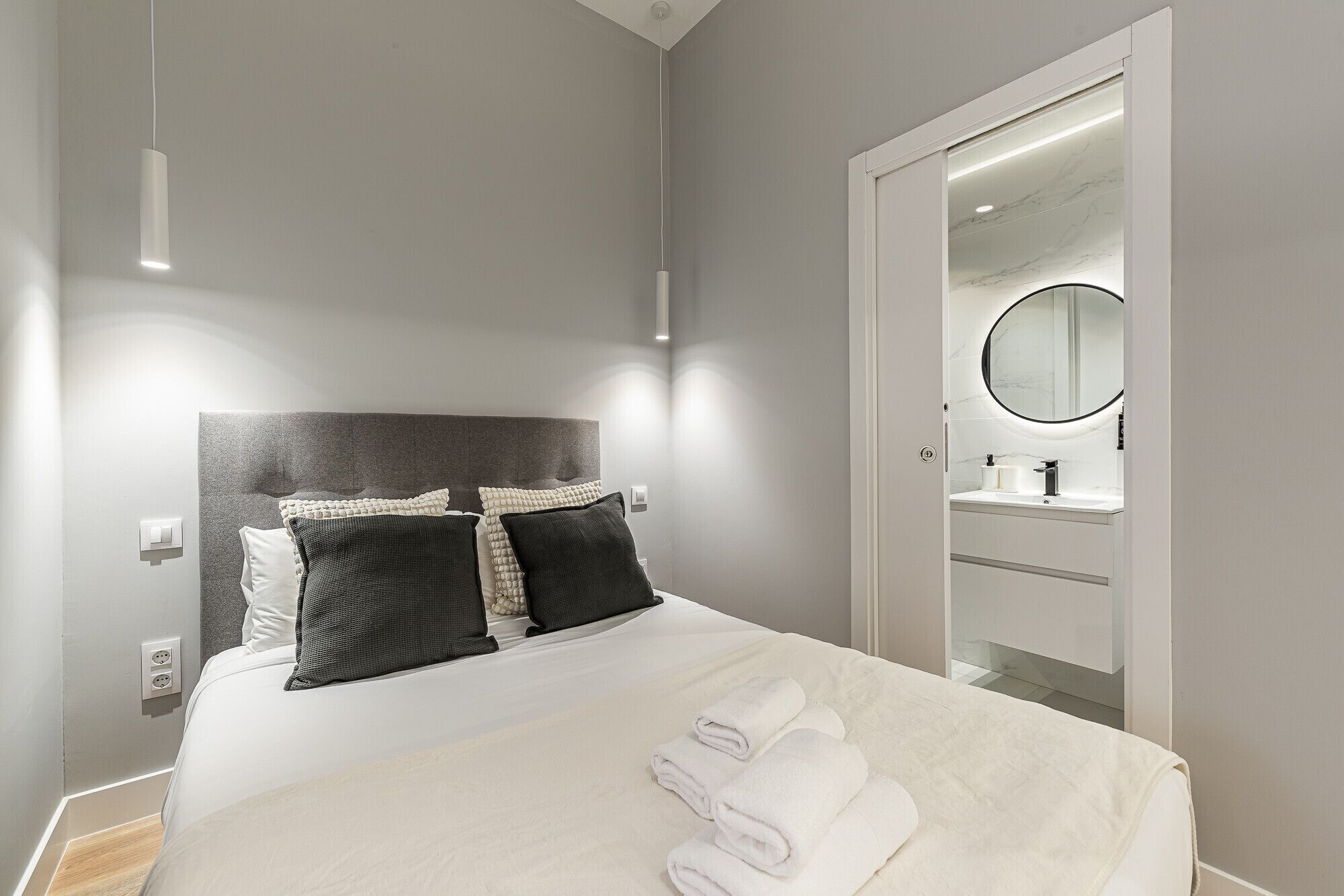 Un dormitorio moderno con cama, almohadas, una mesita de noche y un baño visible a través de una puerta abierta.