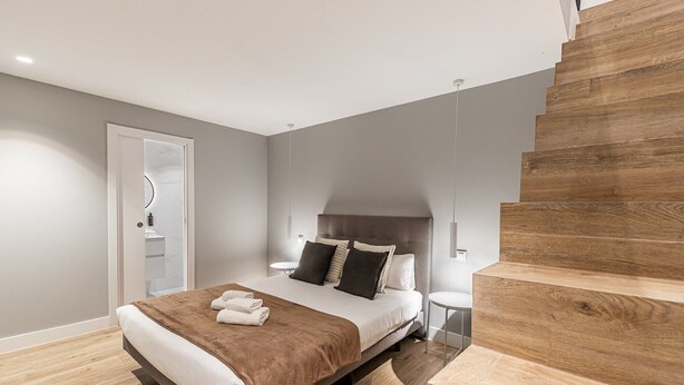 A modern bedroom with a large bed, a wooden staircase, and a bathroom visible through an open door. - Waou Essence III Tirso Molina - Corta Duración (Madrid)