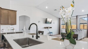 A modern kitchen with a large island, stainless steel sink, and a dining area with a white table and chairs.