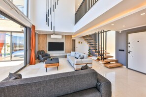 A spacious living area with a modern staircase, a flat-screen TV, and a large window offering a view of the outdoors. - Villa Maranga in Kalkan (Kaş)