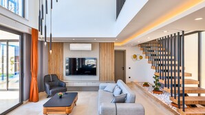 A spacious living area with a large sofa, a coffee table, and a TV. There is a staircase with wooden railings and a view of the outdoors. - Villa Maranga in Kalkan (Kaş)