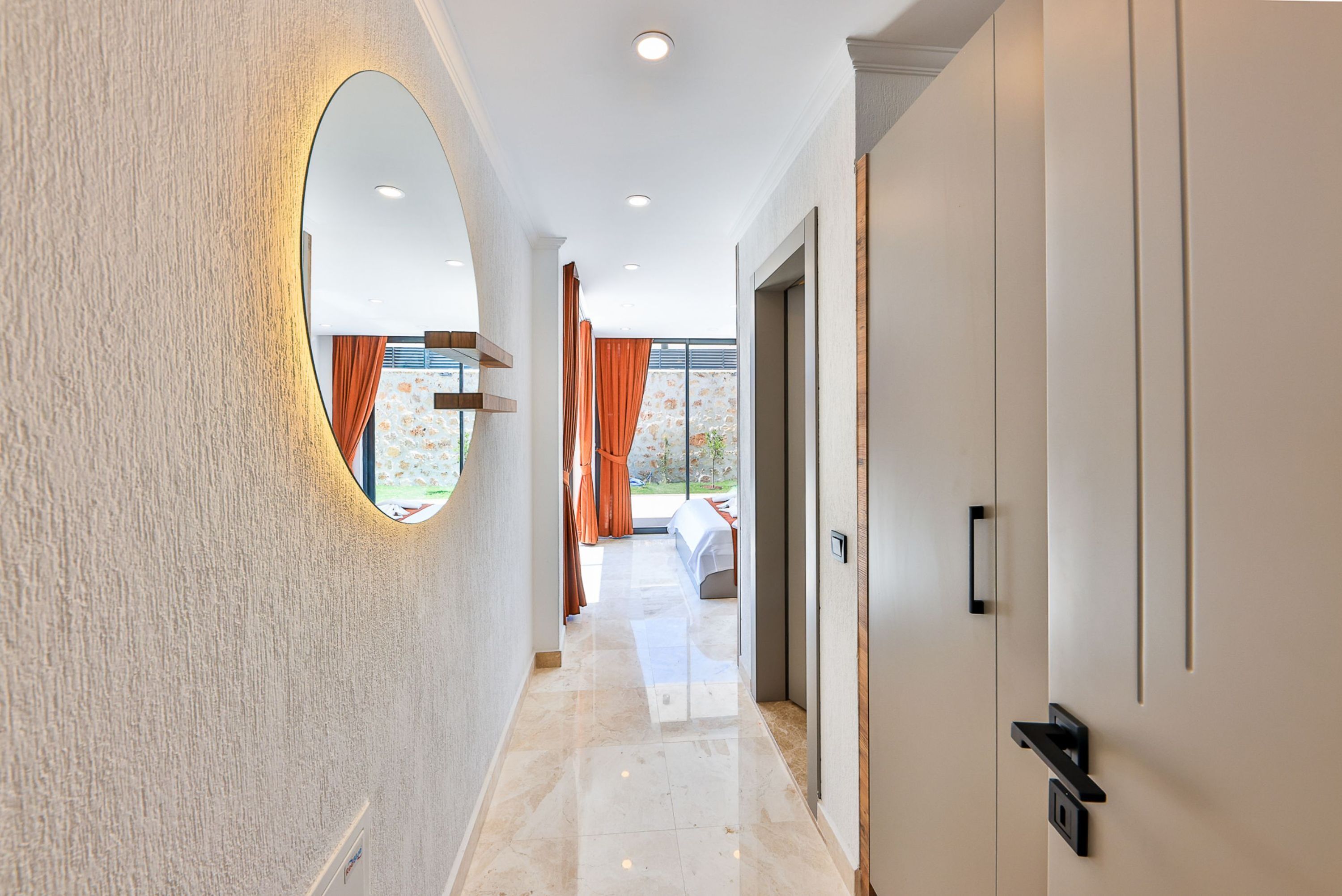 A hallway with a round mirror, a door with a handle, and a view of a patio with a table and chairs.
