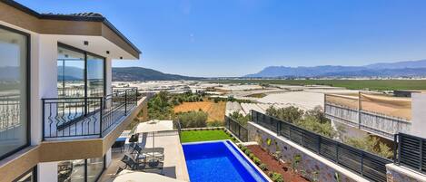 A modern house with a large balcony, a swimming pool, and a view of distant mountains and fields.