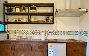Una cocina con armarios de madera, un lavamanos de acero inoxidable y un refrigerador. Hay repisas sobre el lavamanos con varios utensilios de cocina y una pequeña pizarra con un mensaje.
