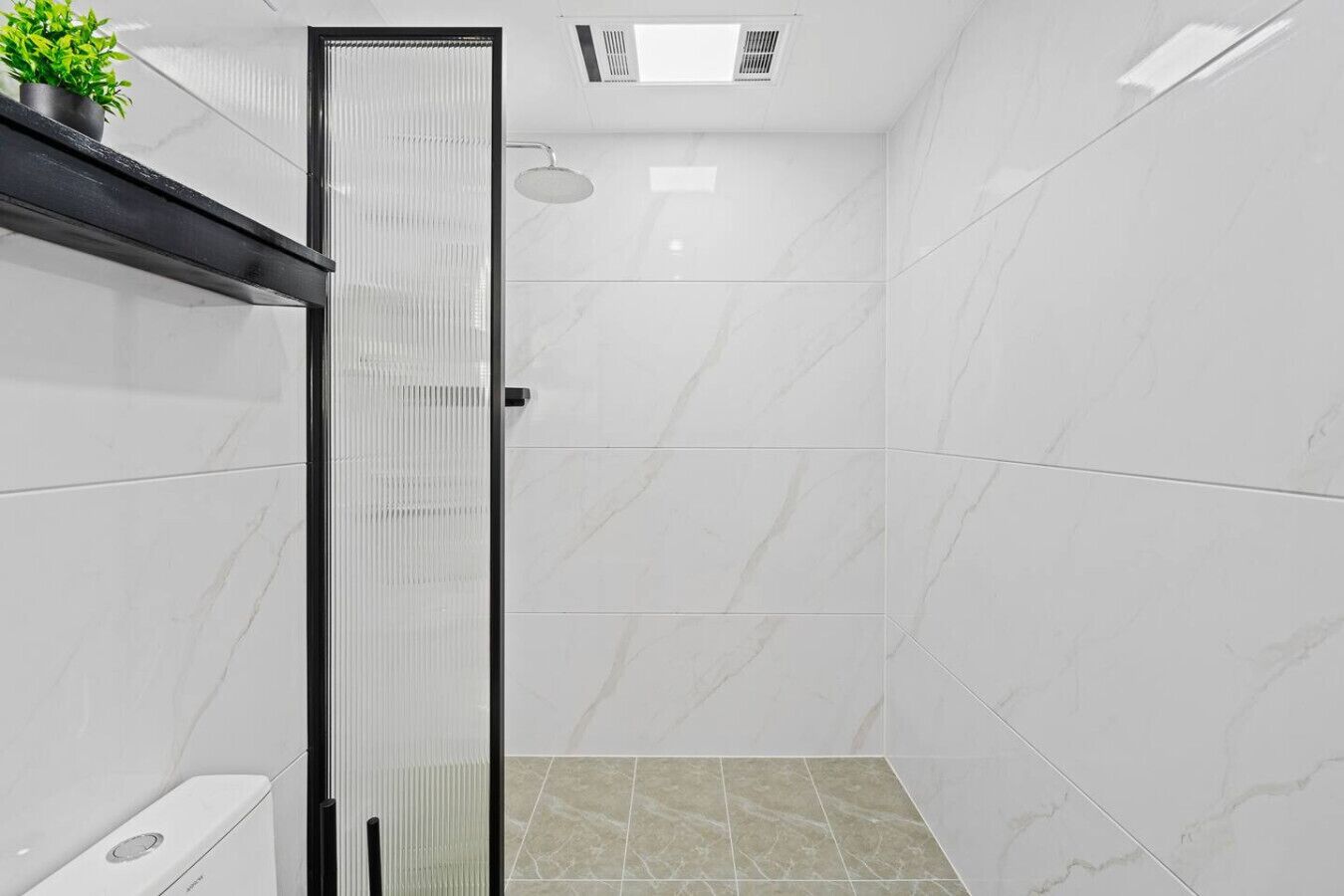 A modern bathroom with a glass shower enclosure, white marble tiles, and a potted plant on the left.