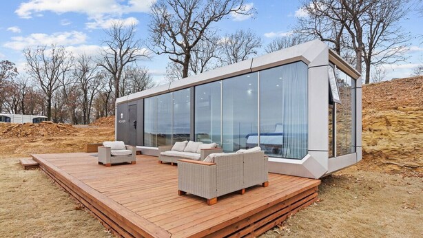 A modern glass house with a wooden deck and outdoor furniture.