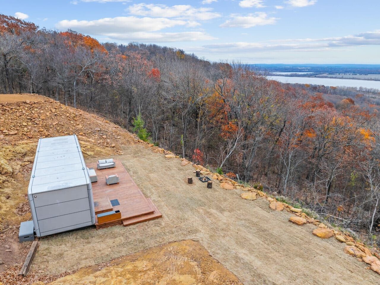 A cabin with a metal roof and a wooden deck, surrounded by a forest with autumn foliage.