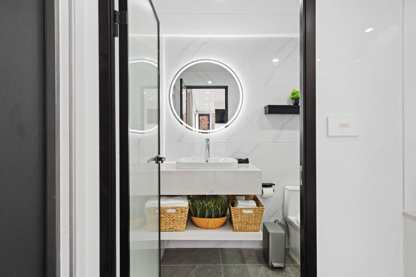 Un bany modern amb porta de vidre, un lavabo blanc, un mirall rodó i una planta petita.