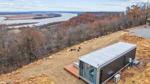 A modern, compact cabin with a glass door and a flat roof, situated on a dirt ground with a view of a river and autumn foliage in the background.