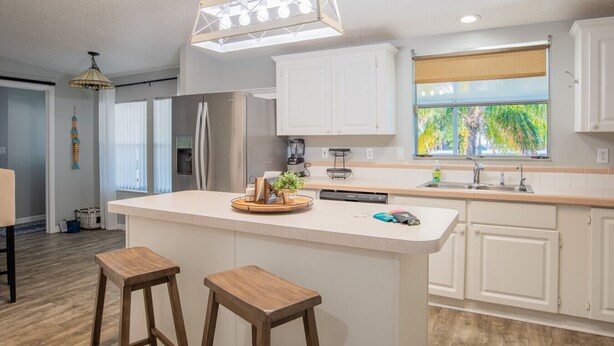 A modern kitchen with white cabinetry, a central island with stools, a built-in dishwasher, and a window with a view of greenery.