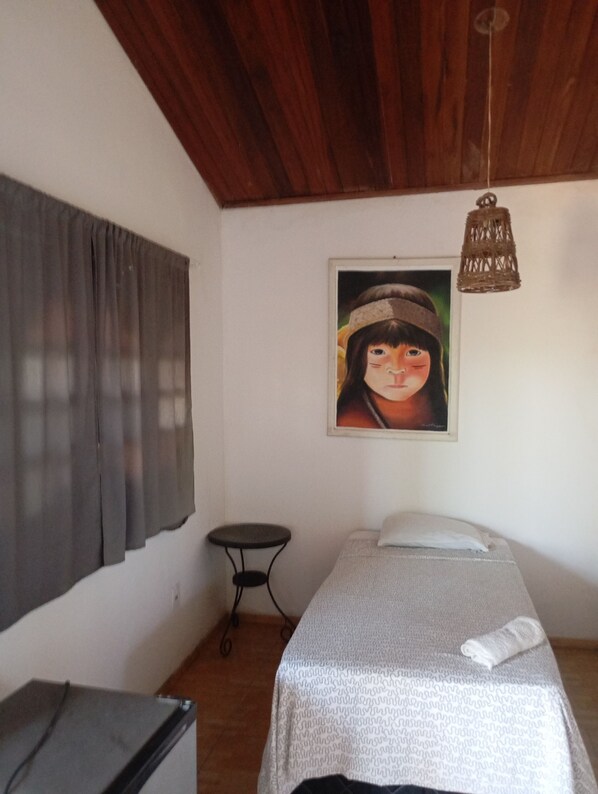A single bed with a white sheet, a small table, a chair, a painting on the wall, and a wooden ceiling. - Guesthouse Paracuru  (Paracuru)