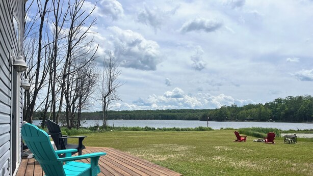 Une terrasse en bois avec deux fauteuils Adirondack turquoise, une petite table et un fauteuil Adirondack rouge. On aperçoit, en arrière-plan, une pelouse verdoyante et un lac bordé d’une rive boisée.