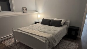A bedroom with a white bed, a patterned rug, a nightstand with a lamp, and a framed picture on the wall. - Relaxing Lake Haven | Peaceful (Saint John)