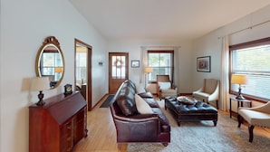 A spacious living room with a leather sofa, a matching ottoman, a wooden sideboard, and a large mirror. There are two chairs, a lamp, and a window with blinds.