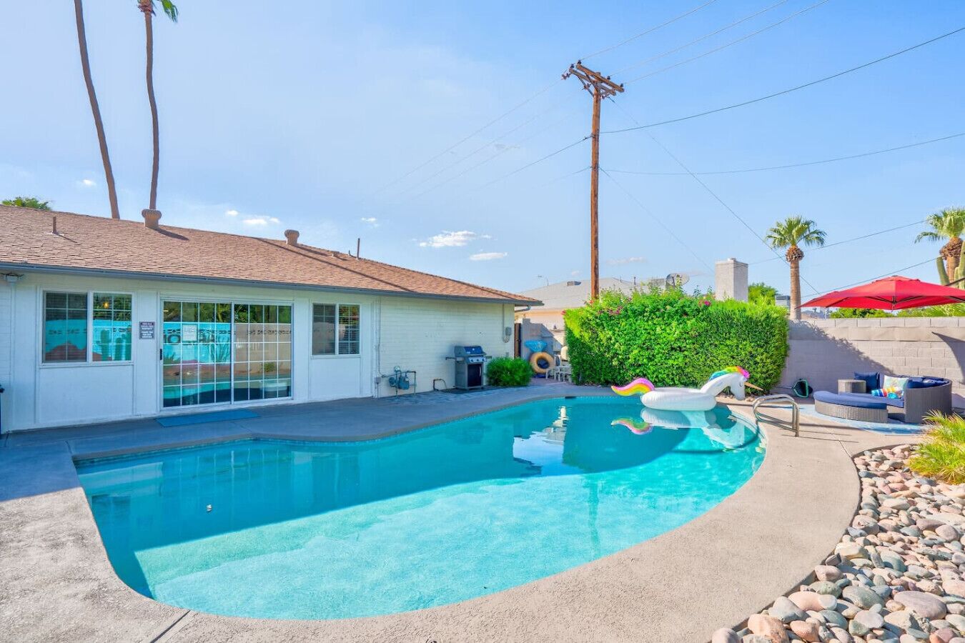 A swimming pool with a clear blue water, surrounded by a concrete deck. There is a small building with a tiled roof and a window.