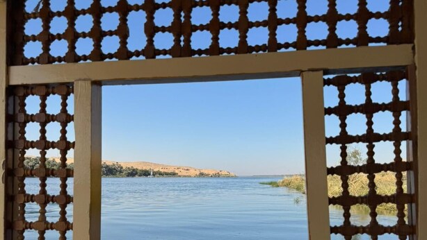 A window with a geometric patterned frame overlooking a calm body of water and distant land.